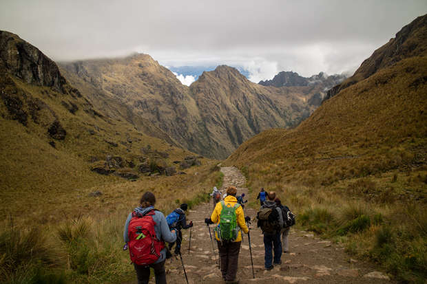 Hikers wearing backpacks walk down a stone path, using trekking poles, surrounded by a mountainous landscape with grassy slopes and a cloudy sky.